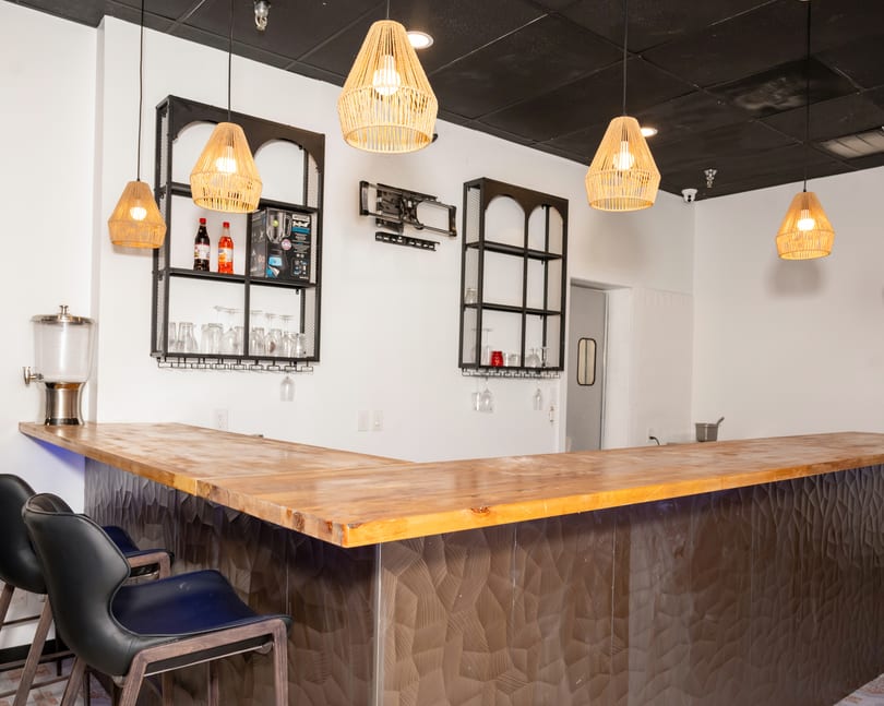 A modern bar area with a wooden countertop, black textured base, and wicker pendant lights.