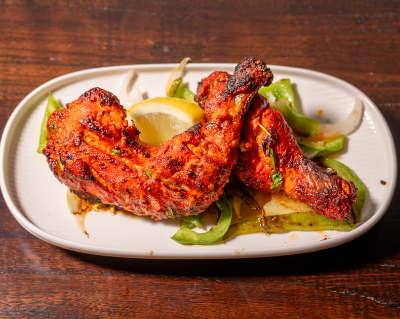 Two vibrant red tandoori chicken legs with charred skin, lemon, and sautéed peppers on a white plate.