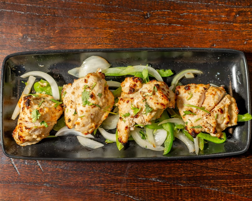 Four pieces of grilled chicken tikka with onions and green peppers on a black plate.