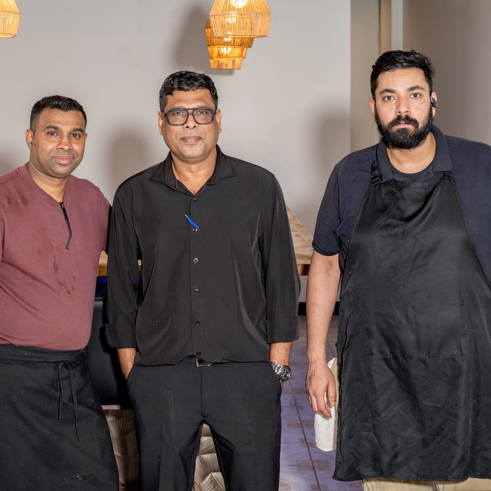Three men, likely restaurant staff, stand together in a dining area with woven light fixtures.