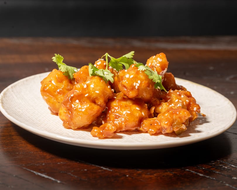 Orange chicken with sesame seeds and cilantro on a white plate, on a dark wooden table.