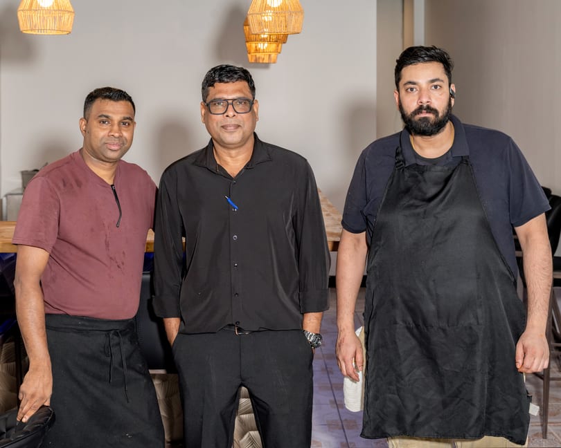 Three men, two in aprons, stand together in a restaurant with woven light fixtures.