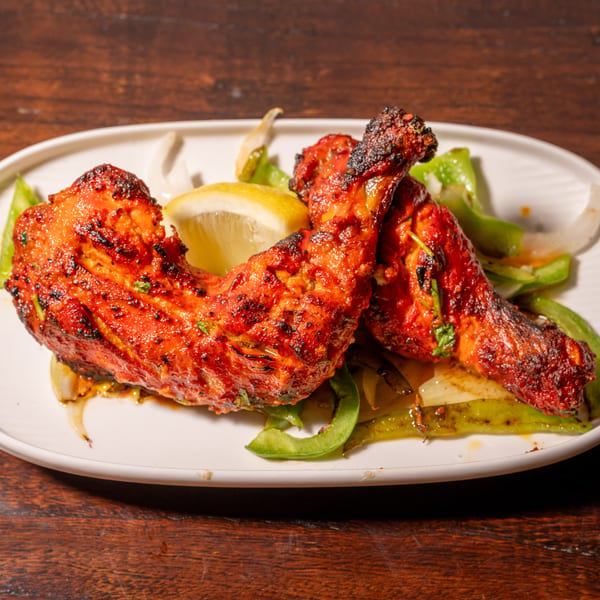 Two vibrant red tandoori chicken legs with charred skin, lemon, and sautéed peppers on a white plate.