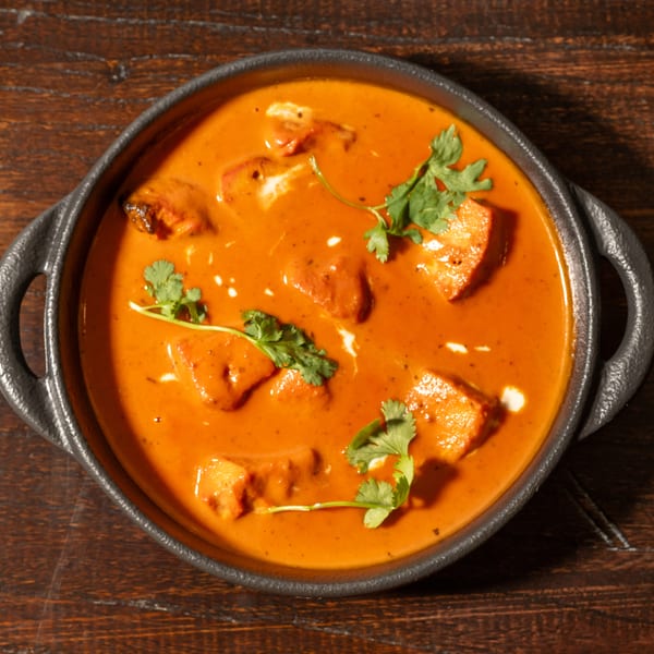 Overhead view of a black bowl filled with orange curry, paneer, and cilantro on a wooden table.
