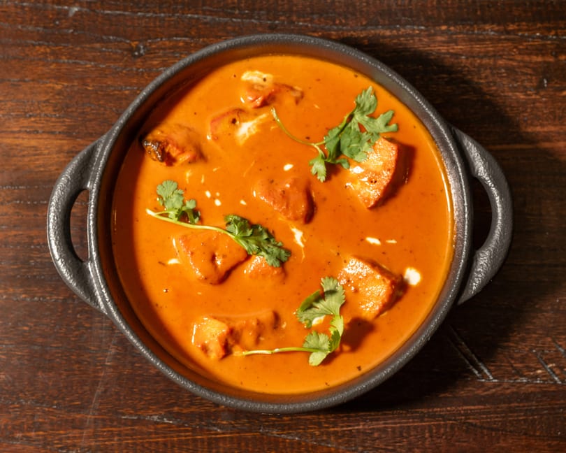 Overhead view of a black bowl filled with orange curry, paneer, and cilantro on a wooden table.