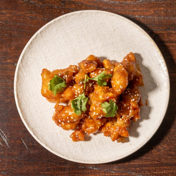 Crispy orange chicken with sesame seeds and cilantro on a white plate, on a wooden table.