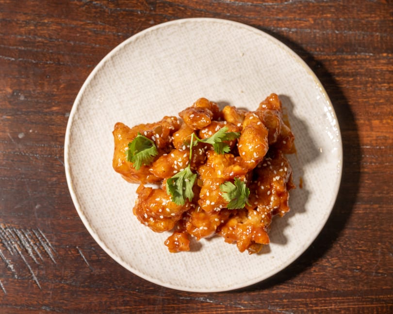 Crispy orange chicken with sesame seeds and cilantro on a white plate, on a wooden table.